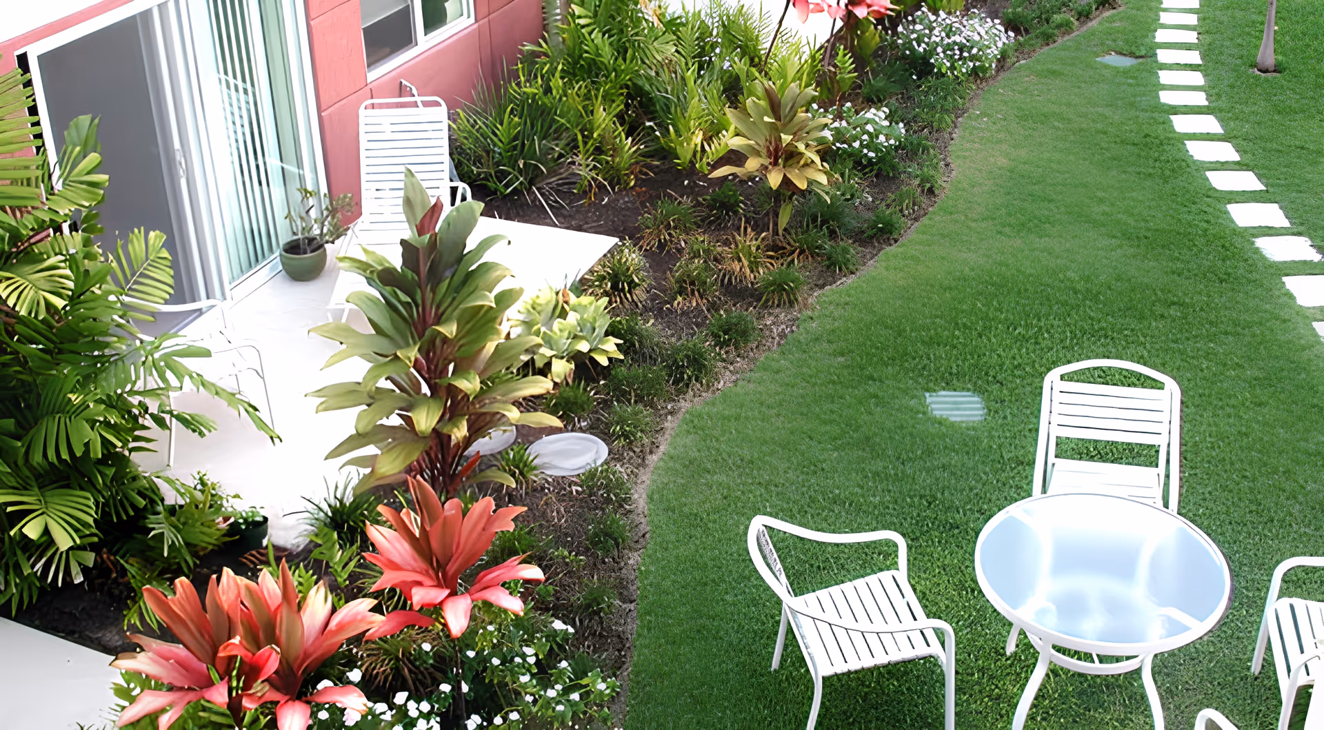 Outdoor garden area with green grass, a curved stone pathway, white metal chairs, and a round white metal table. There are various tropical plants and flowers along the edge of the garden near a building with sliding glass doors and a pink exterior wall.