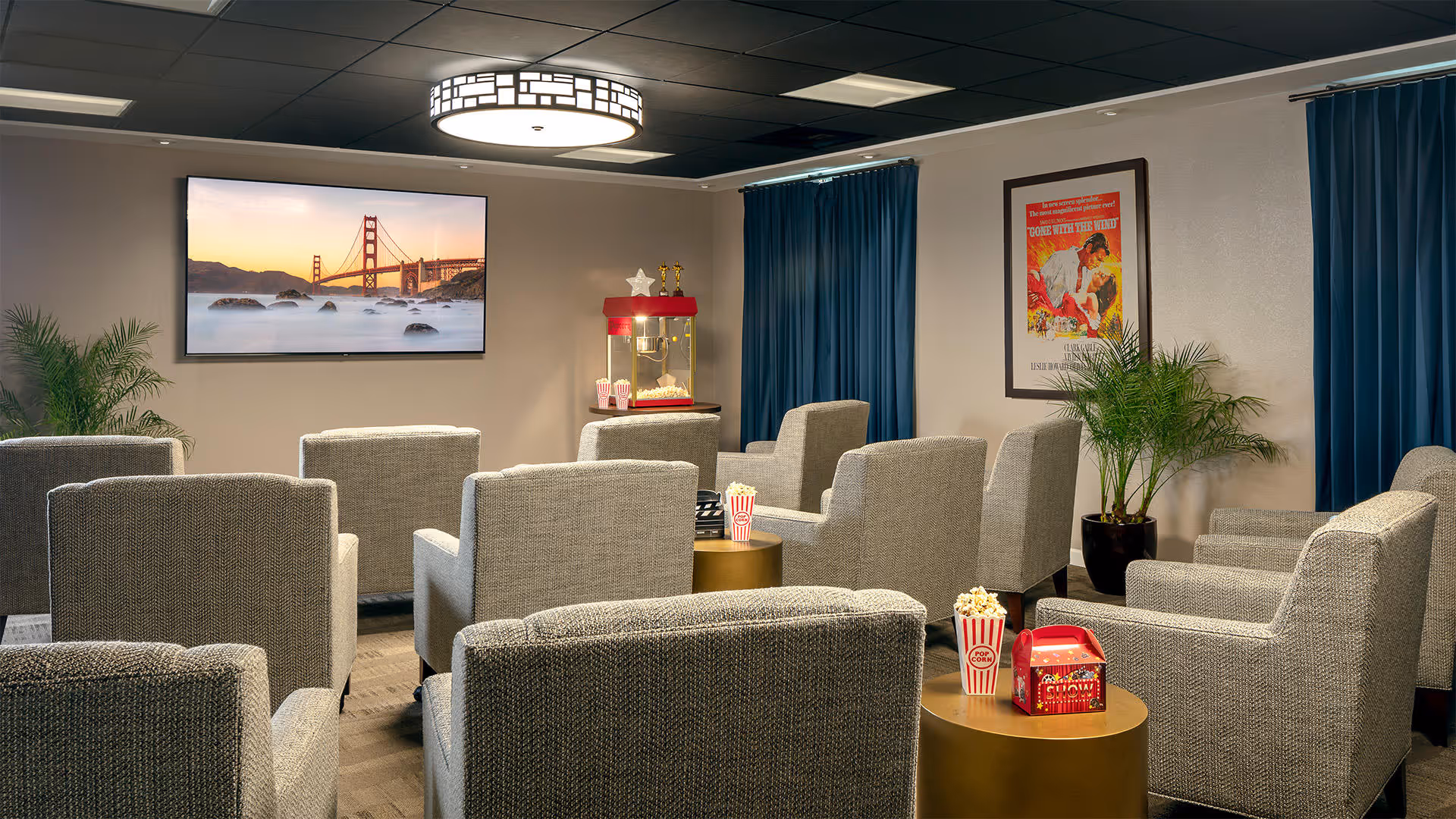 A cozy movie theater room with multiple gray upholstered armchairs arranged in rows facing a large flat-screen TV displaying an image of the Golden Gate Bridge. There is a popcorn machine in the back corner, small round tables with popcorn containers and a red box, blue curtains on the windows, and potted plants adding greenery to the space.