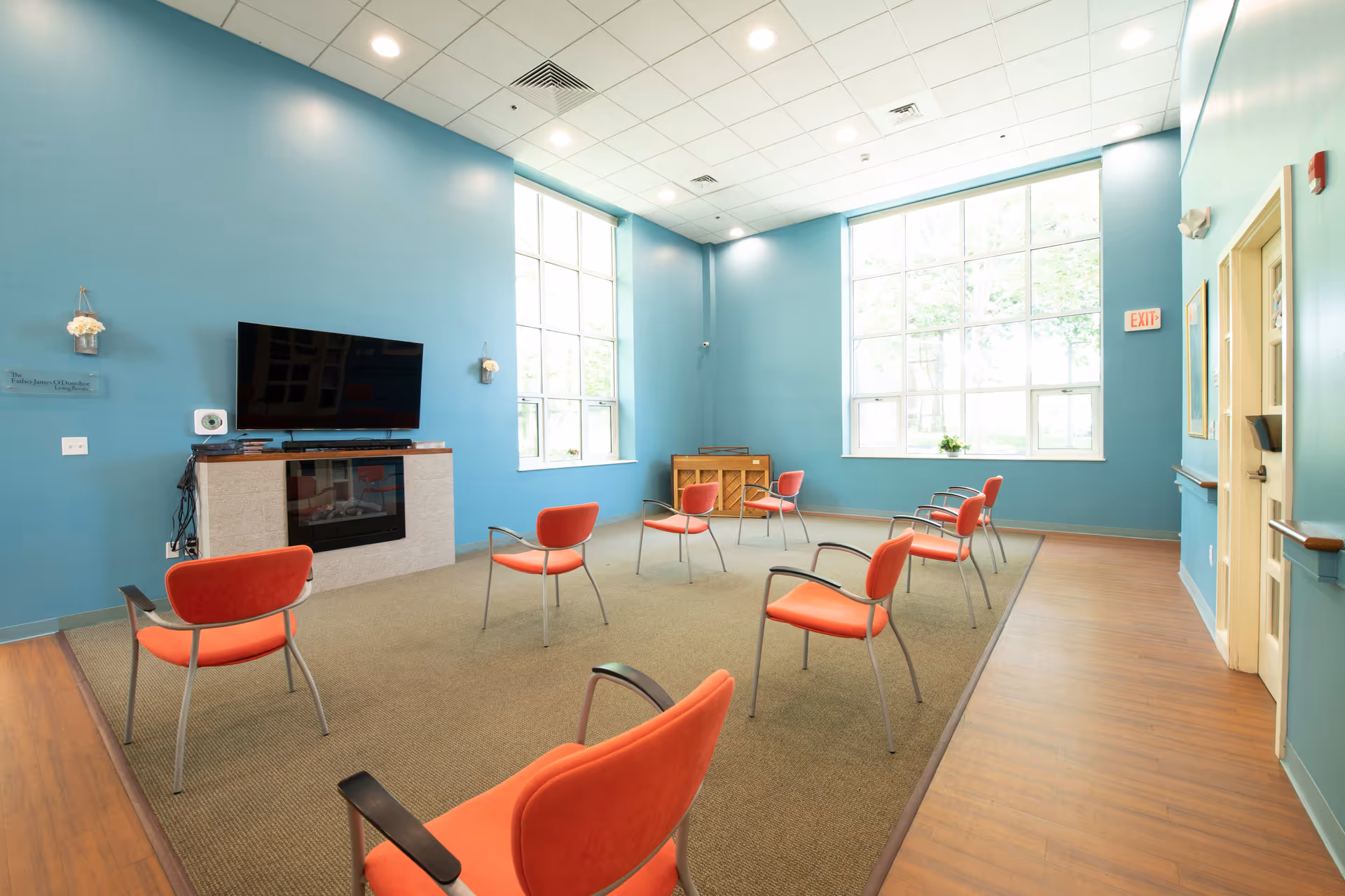 A bright room with blue walls and large windows letting in natural light. The room contains several orange chairs arranged in a spaced-out circle on a carpeted area. A flat-screen TV is mounted on the wall above a fireplace, and a piano is positioned against the far wall. The floor around the carpet is wooden, and there is an exit sign above a door on the right side.