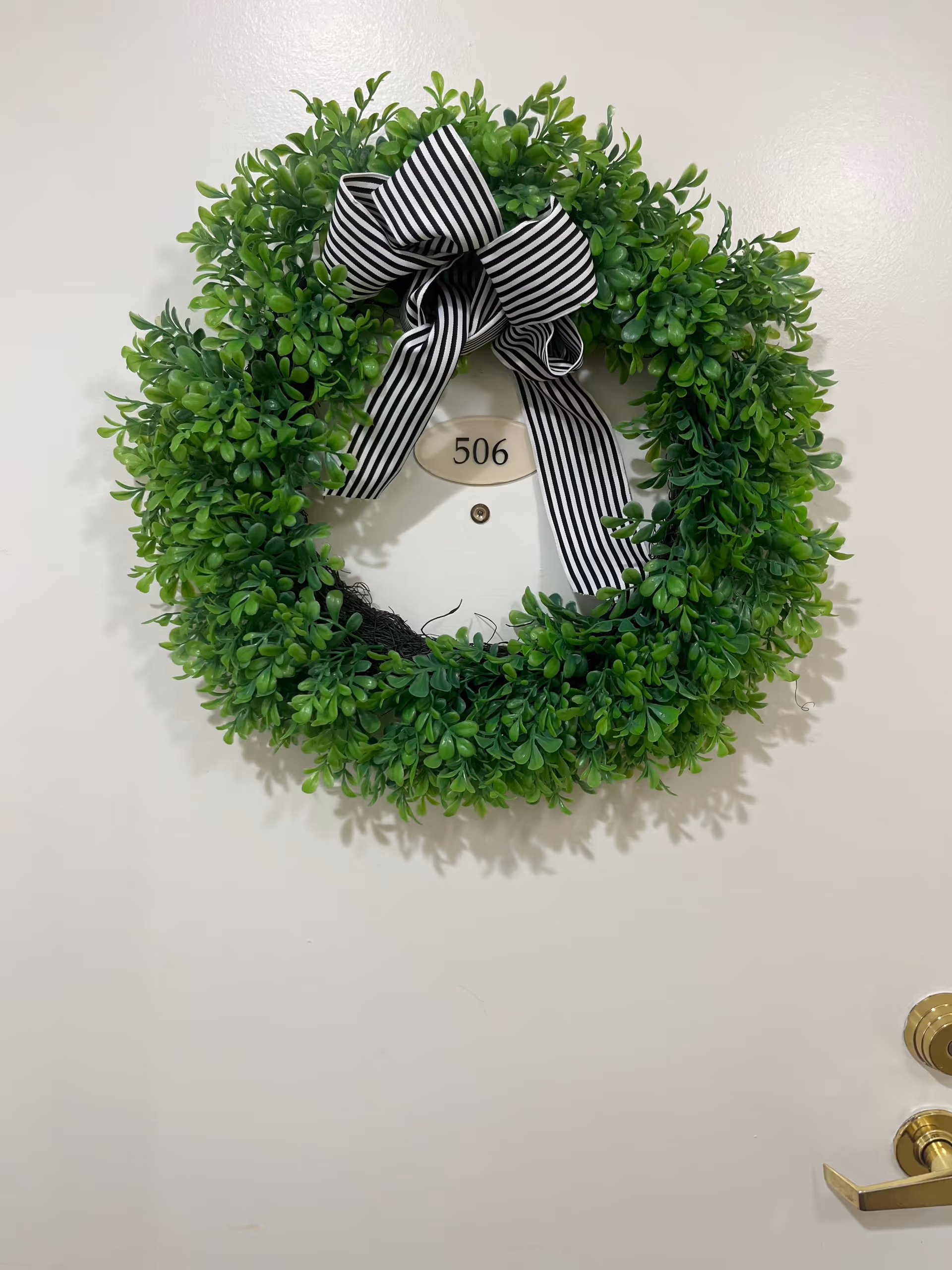 A white door with a green leafy wreath decorated with a black and white striped bow hanging on it. The door has a room number plaque labeled 506 and a peephole below the plaque. A gold-colored door handle and lock are visible on the right side of the door.