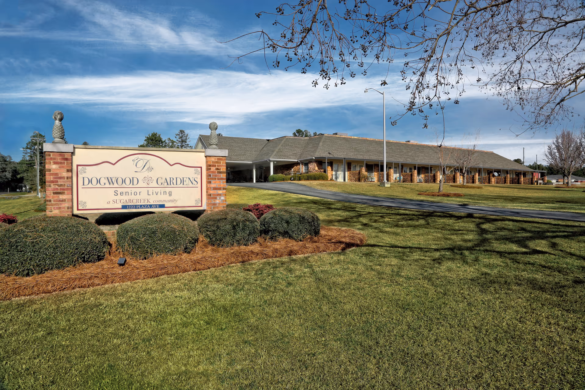 Exterior view of Dogwood Gardens Senior Living facility with a large sign in the foreground displaying the facility name. The building is a single-story structure with a covered entrance, surrounded by well-maintained grass, bushes, and trees under a partly cloudy sky.