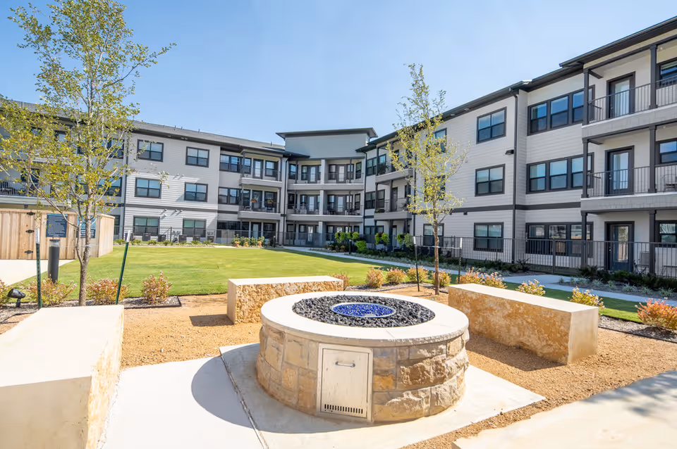 Sunny courtyard with a circular stone fire pit, benches, lawn and a three-story apartment building surrounding it.