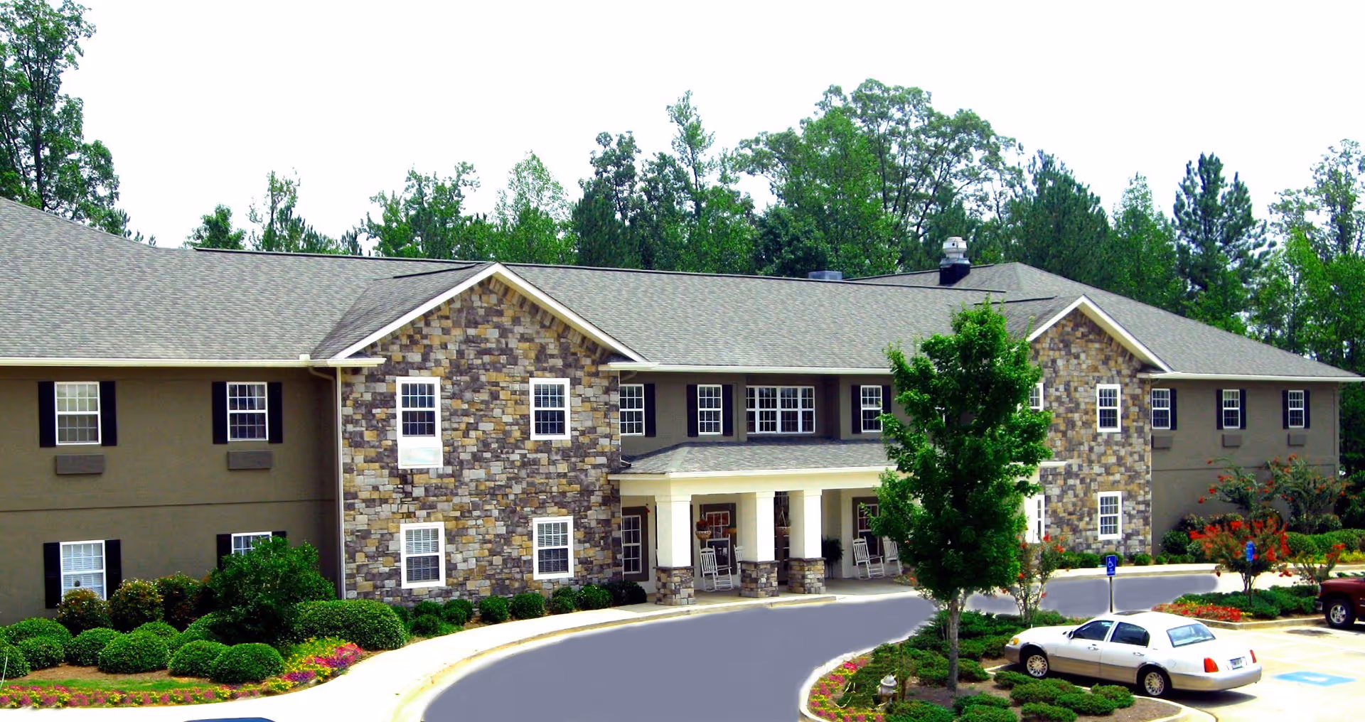Exterior view of Benton Village of Stockbridge, a two-story senior living facility with stone and beige siding, multiple windows with black shutters, a covered entrance with white columns and rocking chairs, surrounded by landscaped bushes, trees, and flowers, with a parking area in front including a white car and a handicapped parking space.