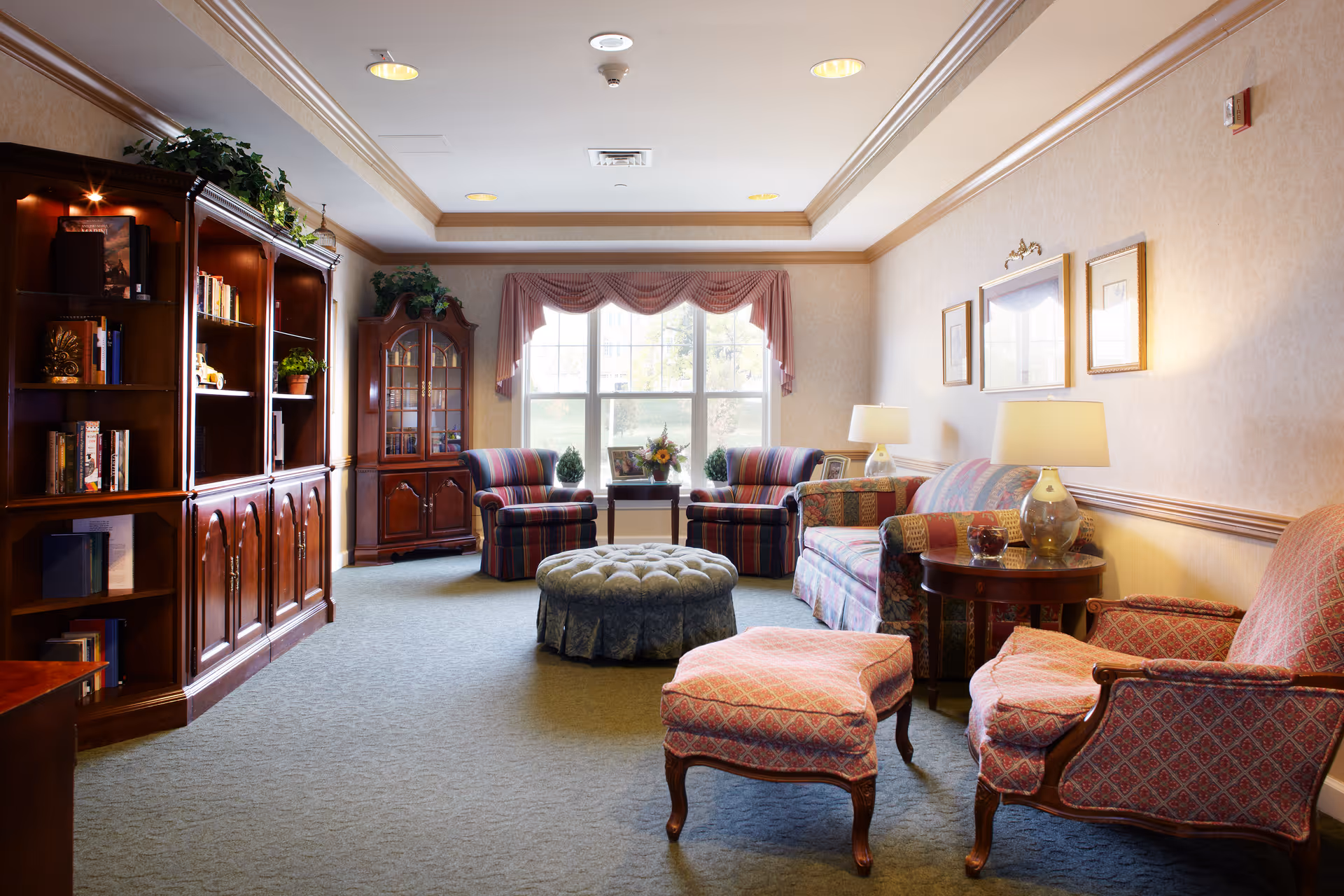 A bright, traditional lounge with patterned armchairs, a sofa, ottomans, wooden bookcases and side tables arranged around a large window.