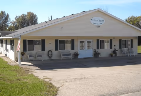 Single-story building with beige siding and a gable roof, featuring a sign that reads 'Country Terrace' above the double front doors. There are several windows with black shutters, benches, potted plants, and an American flag on the left side. The building is surrounded by a paved driveway and green grass under a clear blue sky.
