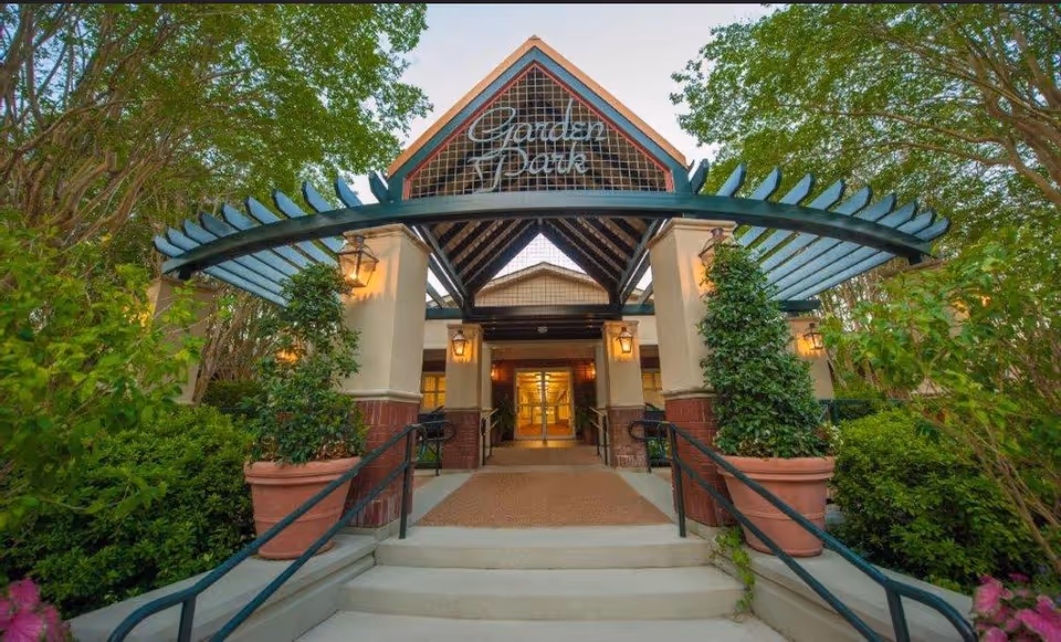 Entrance to Garden Park Nursing & Rehabilitation Center, LLC, featuring a covered walkway with a pergola-style roof, potted plants on either side of the steps, and greenery surrounding the entrance.