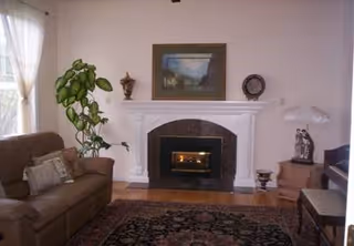 A cozy living room featuring a brown sofa with cushions on the left, a tall green plant next to a window with sheer white curtains, a white fireplace with a dark stone surround in the center, a framed painting above the fireplace, and decorative items on the mantel. To the right, there is a wooden side table with a lamp and a figurine, a piano with a bench, and a patterned area rug covering the wooden floor.