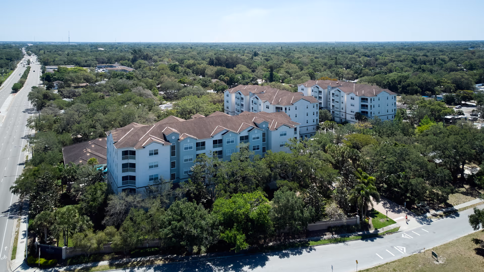 Aerial view of a large senior living facility named Arietta surrounded by dense green trees and vegetation. The facility consists of multiple connected white buildings with brown roofs. A wide road runs alongside the property, and there are sidewalks and smaller roads around the complex.