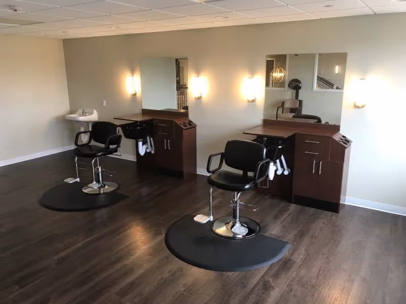 Interior salon area with two black styling chairs at mirrored stations, cabinetry, sinks and wall sconces on a wood floor.