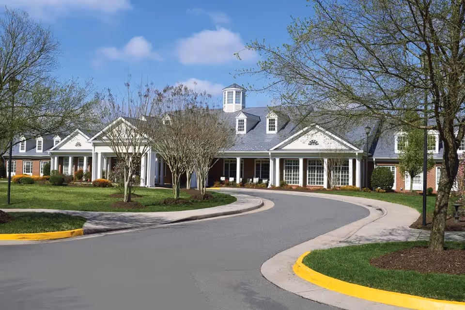 Exterior view of a senior living facility building with a curved driveway, manicured lawns, and trees under a blue sky with some clouds.