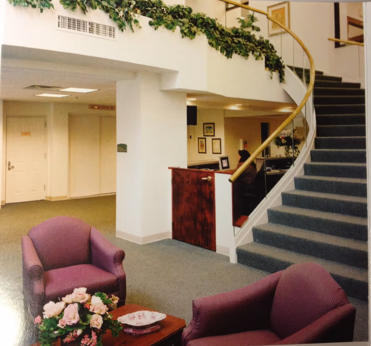 Interior view of a senior living facility lobby area with two purple armchairs and a small wooden table with a floral arrangement and a decorative plate. A curved staircase with a wooden handrail and glass panels leads to an upper floor. A reception desk with a person seated behind it is visible in the background, along with framed artwork on the walls and green garland decoration along the upper ledge.