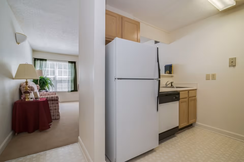 View of a small kitchen area with a white refrigerator, wooden cabinets, a sink, and a dishwasher. Adjacent to the kitchen is a living room with a window covered by green curtains, a plaid armchair, a side table with a red tablecloth, a lamp, and a potted plant.