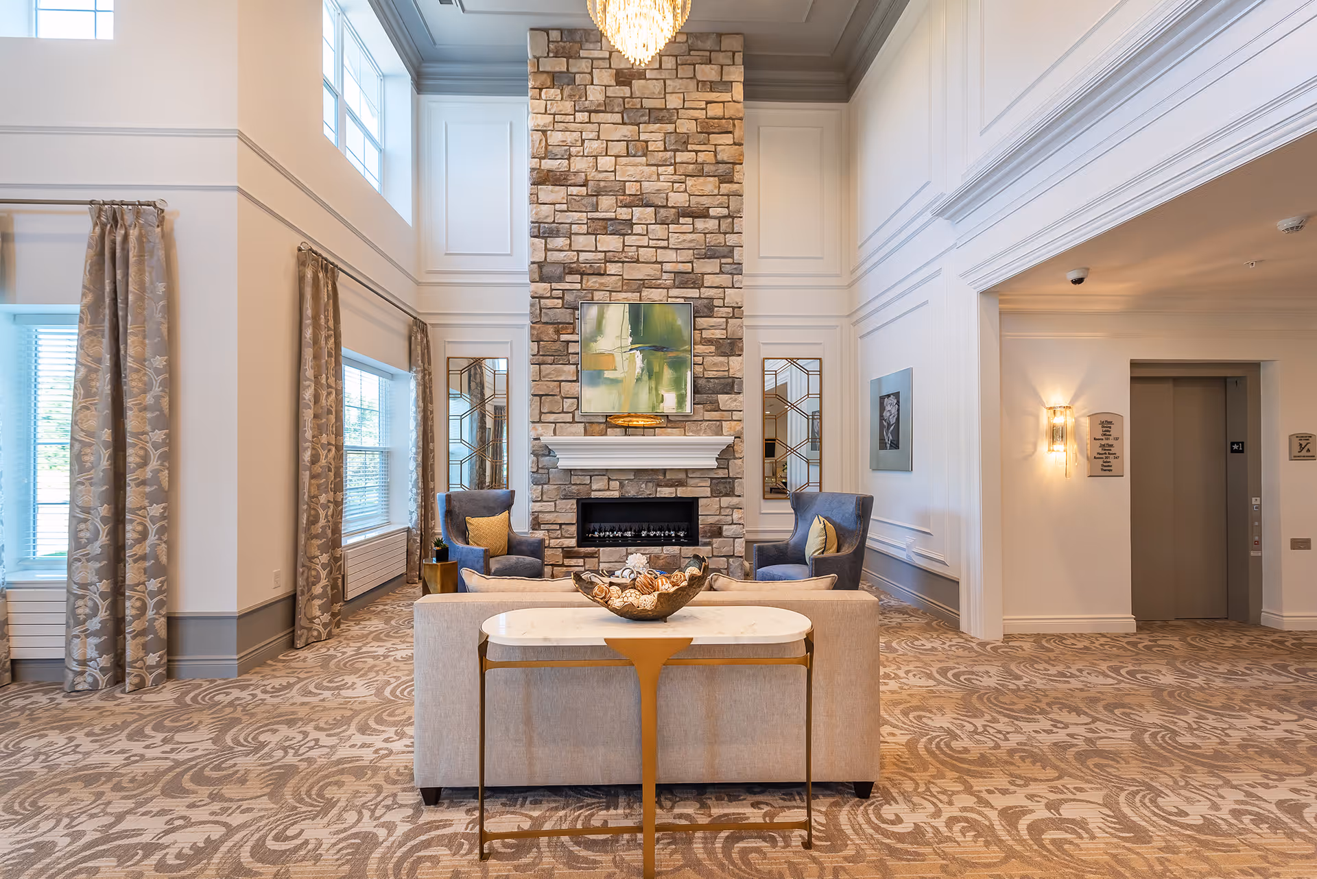 A spacious and elegant living room area with a tall stone fireplace as the focal point. The room features two blue armchairs with yellow pillows, a beige sofa, a small table with decorative items, large windows with patterned curtains, and a chandelier hanging from the ceiling. The walls are white with decorative molding, and there is an elevator visible in the background.
