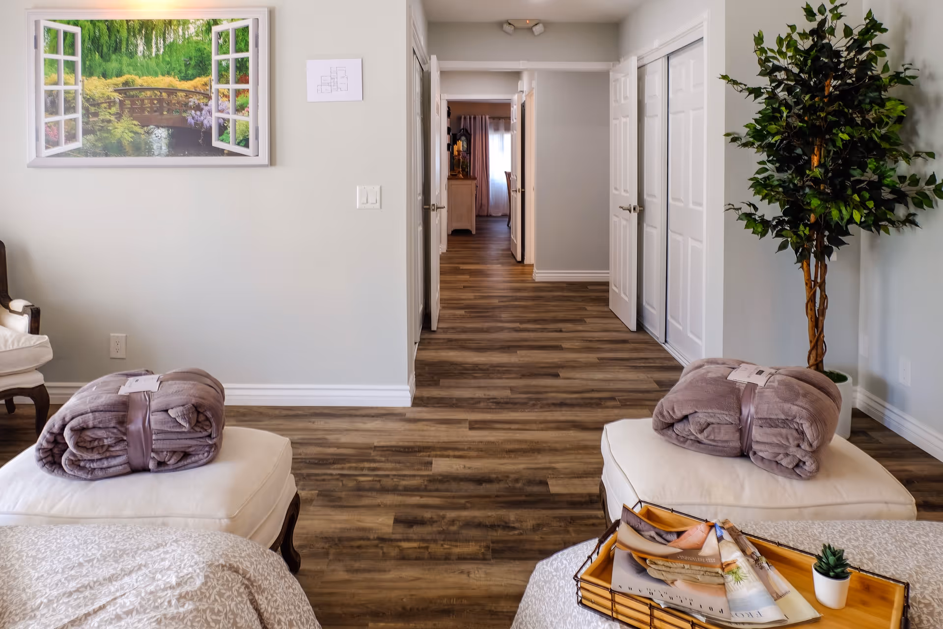 Bright interior bedroom/sitting area with ottomans topped with folded blankets, a tray with magazines, a potted plant, wall art, and a hallway leading to other rooms.