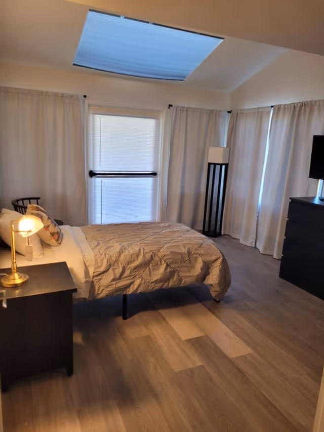 A bedroom with a single bed covered with a beige comforter and a pillow with a floral design. There is a black nightstand with a brass lamp on the left side of the bed. The room has light wood flooring, white curtains covering the windows, and a skylight on the ceiling. A black dresser with a TV on top is visible on the right side, and a tall floor lamp stands in the corner.