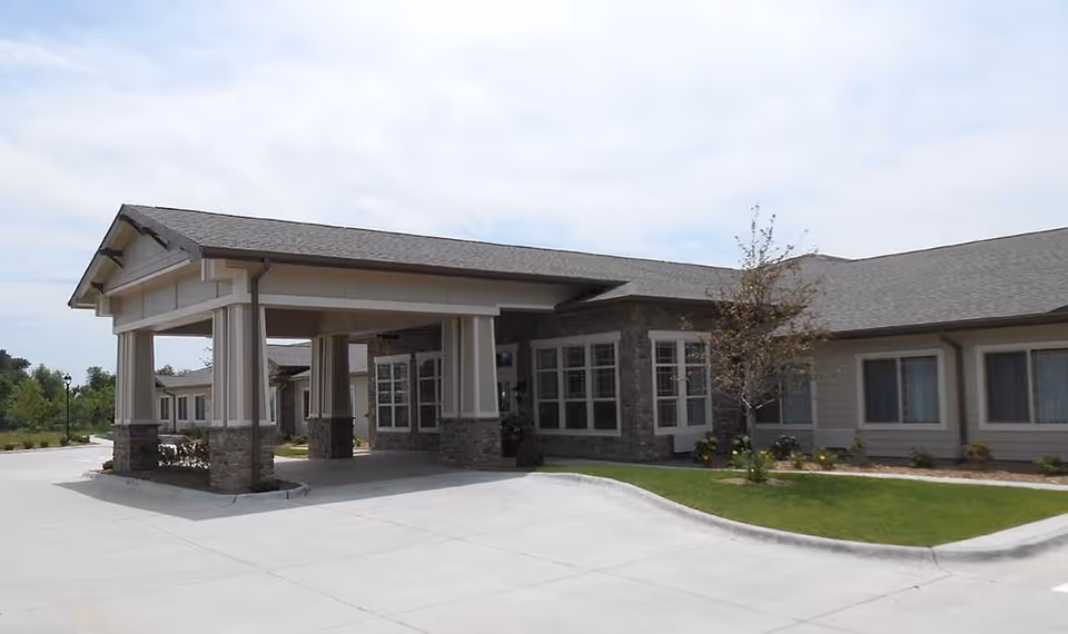 Front entrance of a one-story senior living building with a covered porte-cochere, driveway, and landscaping.