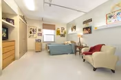 A bright nursing facility bedroom with a single bed, armchair, dresser, TV stand, and wall decorations.