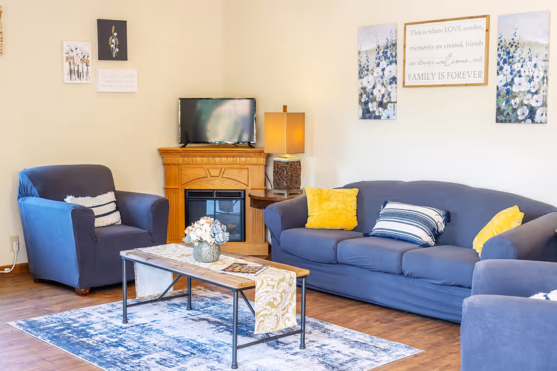 A cozy living room with a blue sofa and armchairs, decorated with yellow and striped pillows. A wooden coffee table with a floral centerpiece and a decorative runner sits on a blue patterned rug. A small wooden electric fireplace with a TV on top and a table lamp is against the wall. The walls are adorned with floral artwork and a framed sign with an inspirational family quote.