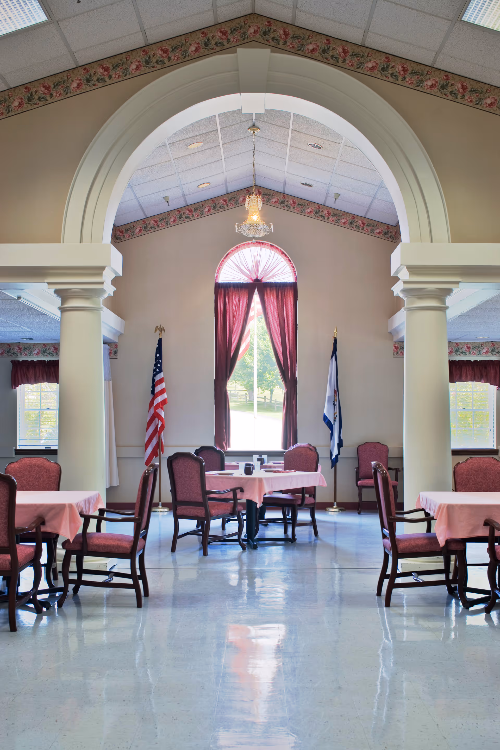 Spacious dining room with an arched entry, tables covered in pink tablecloths, upholstered chairs, a chandelier, and flags flanking a tall window with maroon curtains.