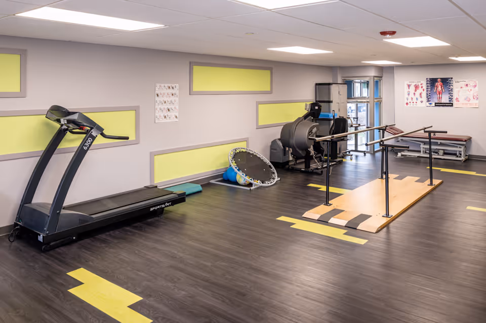 A rehabilitation exercise room with a treadmill, a small trampoline, exercise balls, parallel bars for walking practice, and medical posters on the walls. The room has dark wood flooring with yellow markings and light gray walls with yellow accent panels.