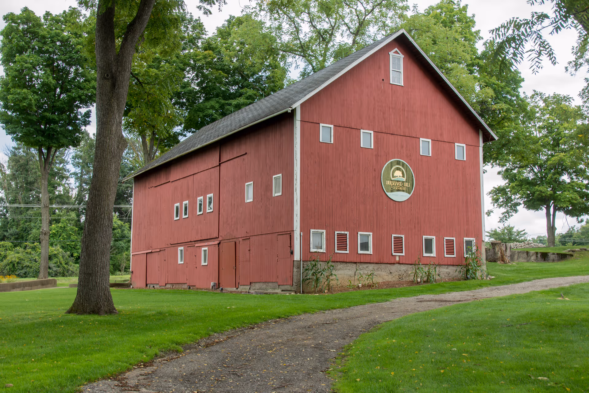 A large red barn-style building with multiple small windows and a sign that reads 'Hubbard Hill' on the side, surrounded by green grass and trees under a cloudy sky.