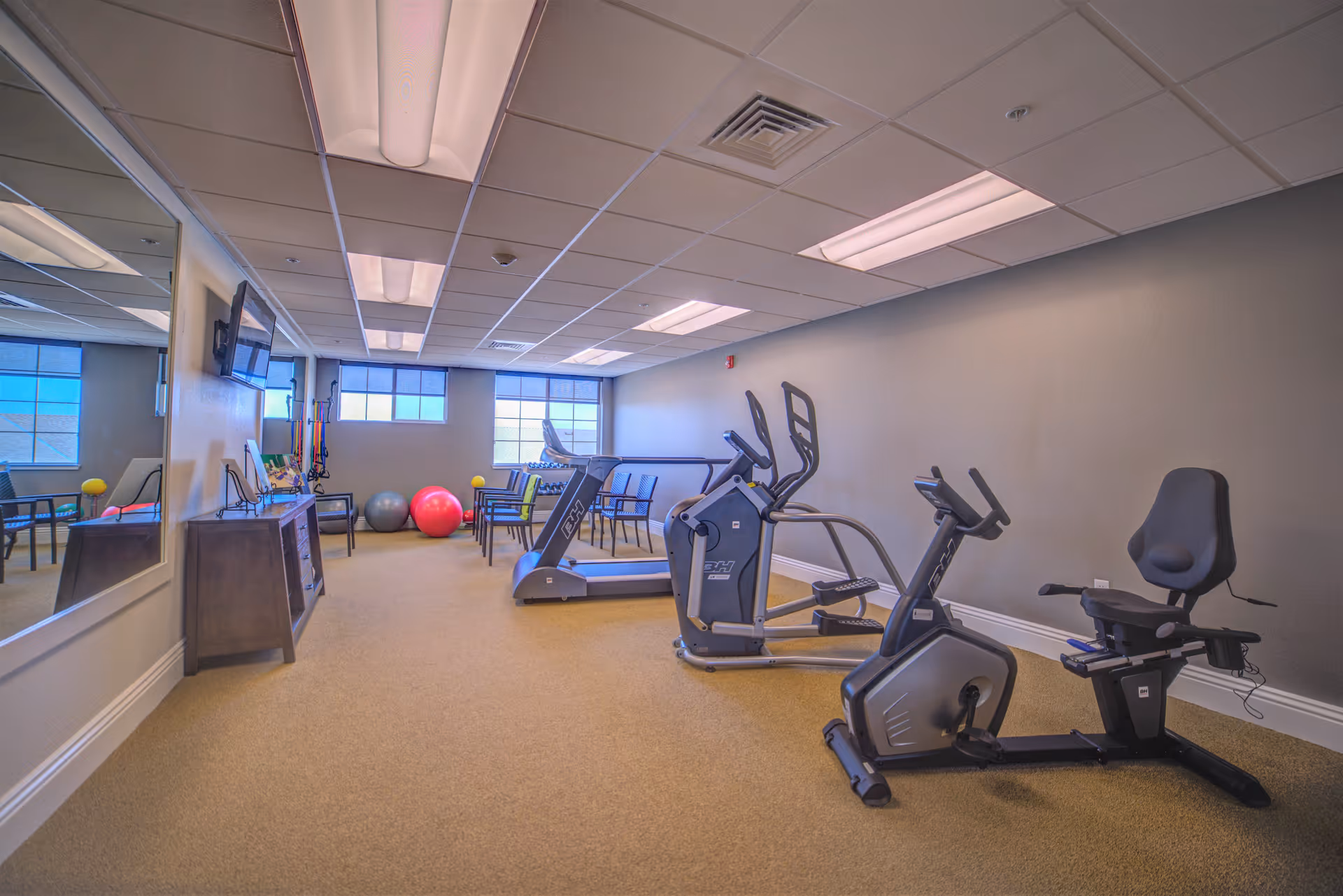 A fitness room with exercise equipment including a recumbent bike, elliptical machine, and treadmill. There are also colorful exercise balls and chairs near the windows. The room has beige carpet, gray walls, and a large mirror on one side.