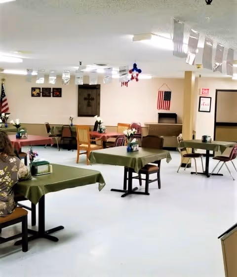 A dining area in a nursing home with several tables covered in green and pink tablecloths, each decorated with small flower arrangements. There are various chairs around the tables, an American flag on the wall, and ceiling decorations hanging down. The room has a light-colored floor and walls, with an exit sign visible in the background.