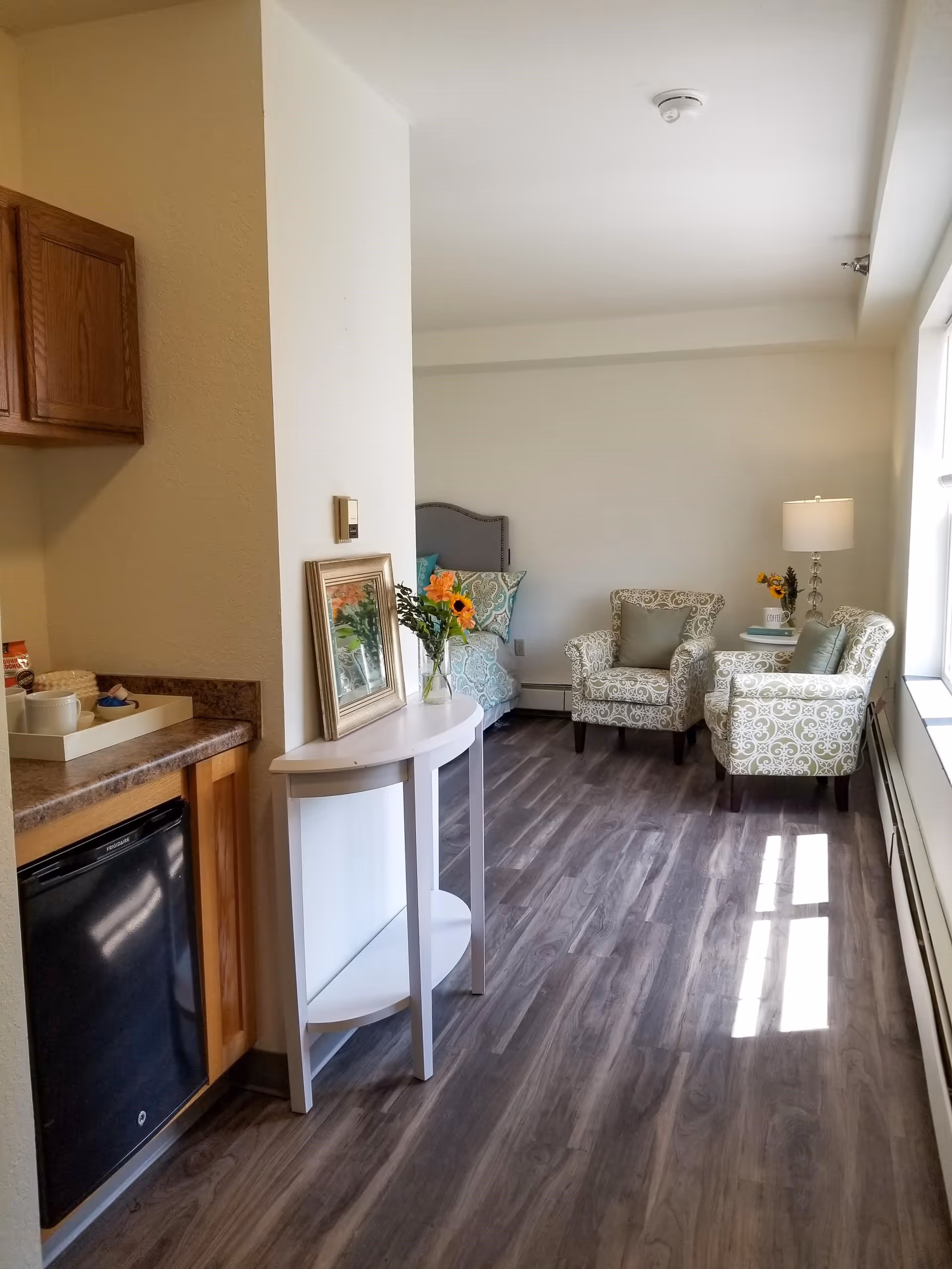 A bright interior room with wood flooring featuring a small kitchenette area with a mini fridge and countertop on the left. A white half-moon table with a framed picture and a vase of sunflowers is placed against a partial wall. In the background, there is a sitting area with two patterned armchairs, a small round table with a lamp and flowers, and a bed with decorative pillows. Sunlight streams in through a window on the right.