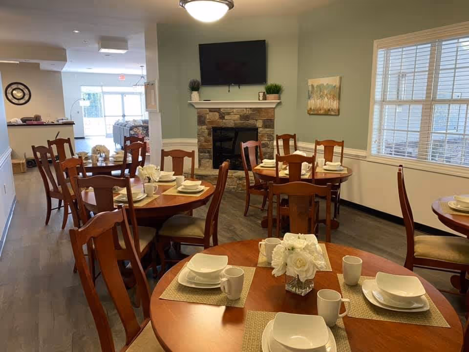 A dining room with several round wooden tables set with white plates, bowls, mugs, and beige placemats. Each table has a small floral centerpiece. The room has wooden chairs, a stone fireplace with a TV mounted above it, a large window with blinds, and light green walls.