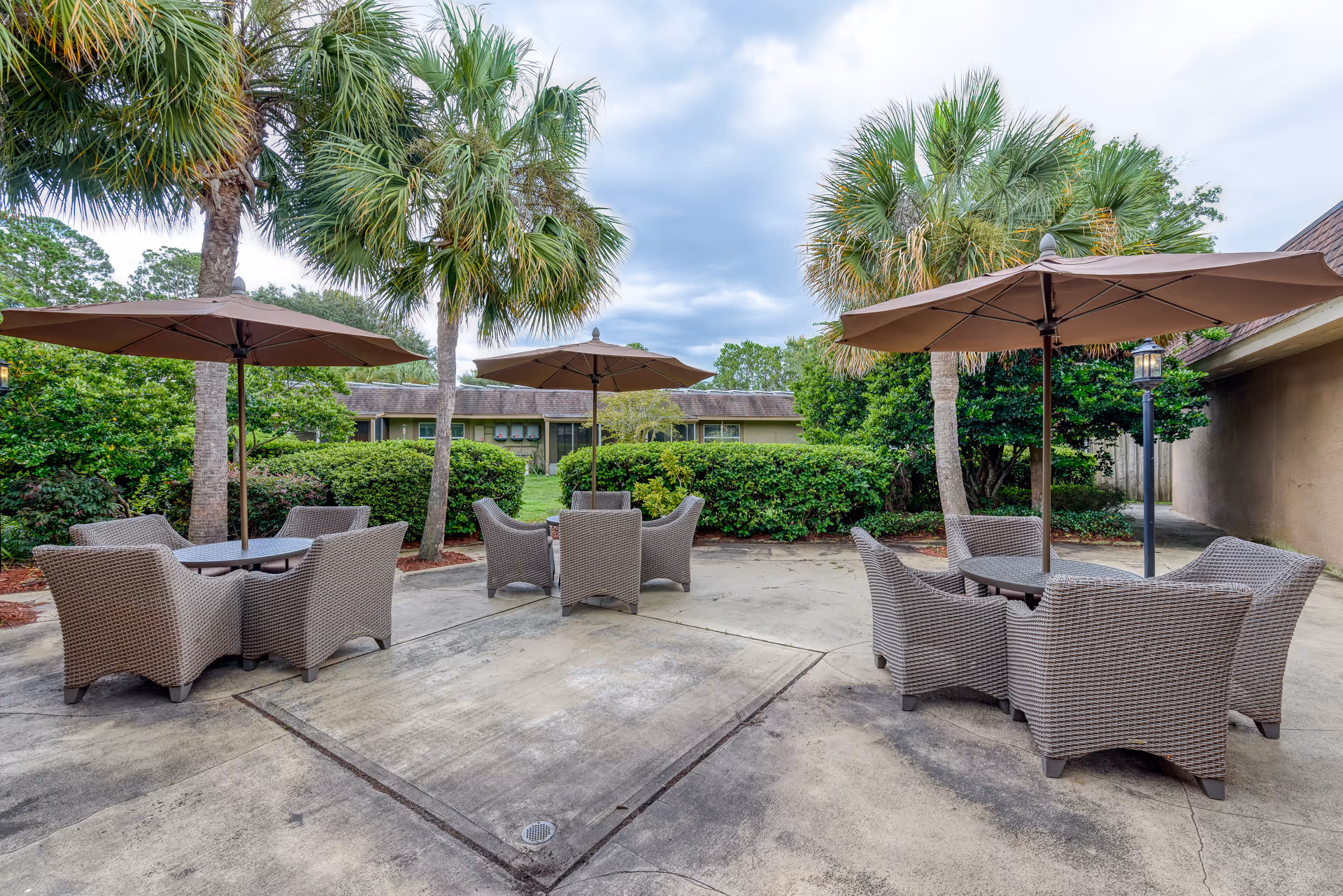 Outdoor patio area with three round tables, each surrounded by four wicker chairs and shaded by large brown umbrellas. The patio is surrounded by palm trees, green bushes, and a building wall on the right side under a cloudy sky.