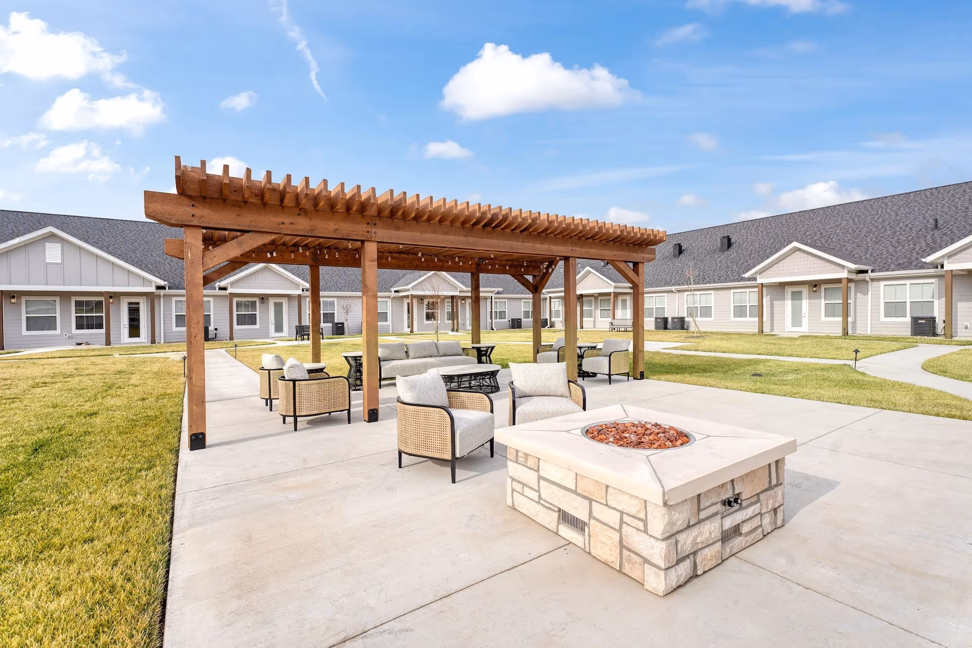 Outdoor seating area at Cedarhurst Senior Living of Salina featuring a wooden pergola with string lights, cushioned chairs, a sofa, and a stone fire pit on a concrete patio surrounded by green grass and single-story residential buildings.