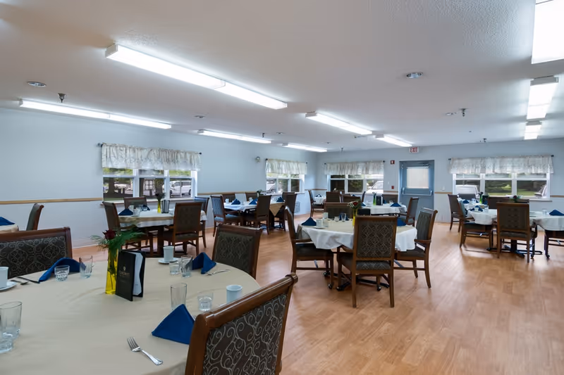 Spacious dining room with multiple round tables set with white tablecloths, blue napkins, and wooden chairs on light wood flooring.