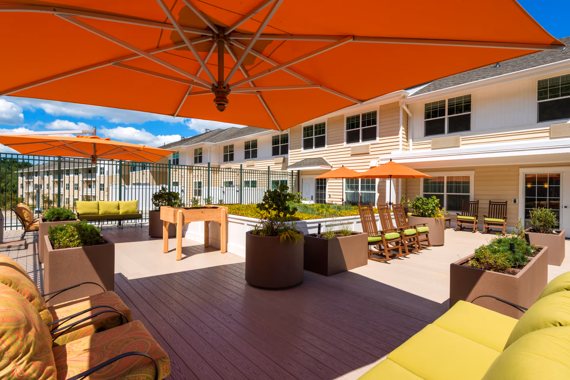 Outdoor patio area at a senior living facility with large orange umbrellas providing shade over seating areas. There are cushioned chairs, rocking chairs, and planters with greenery. The building with multiple windows is visible in the background under a blue sky with some clouds.