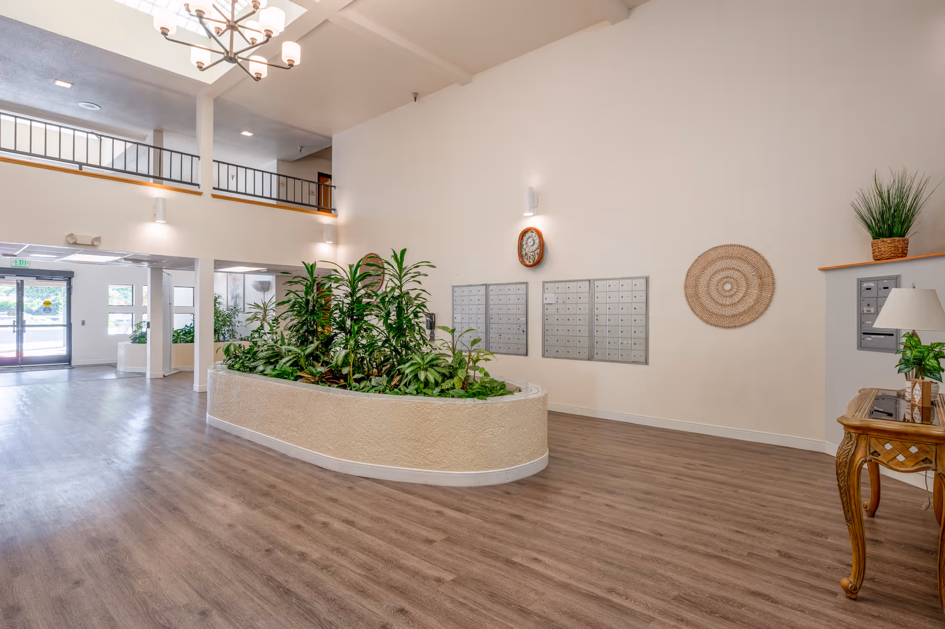 Spacious and well-lit lobby area with a large planter filled with green plants in the center. There are multiple mailboxes mounted on the wall, a decorative wall clock, and a woven wall hanging. The floor is wood-style laminate, and there is a wooden table with a lamp and a potted plant on the right side. The ceiling is high with a chandelier, and there is a second-floor railing visible above the entrance area.