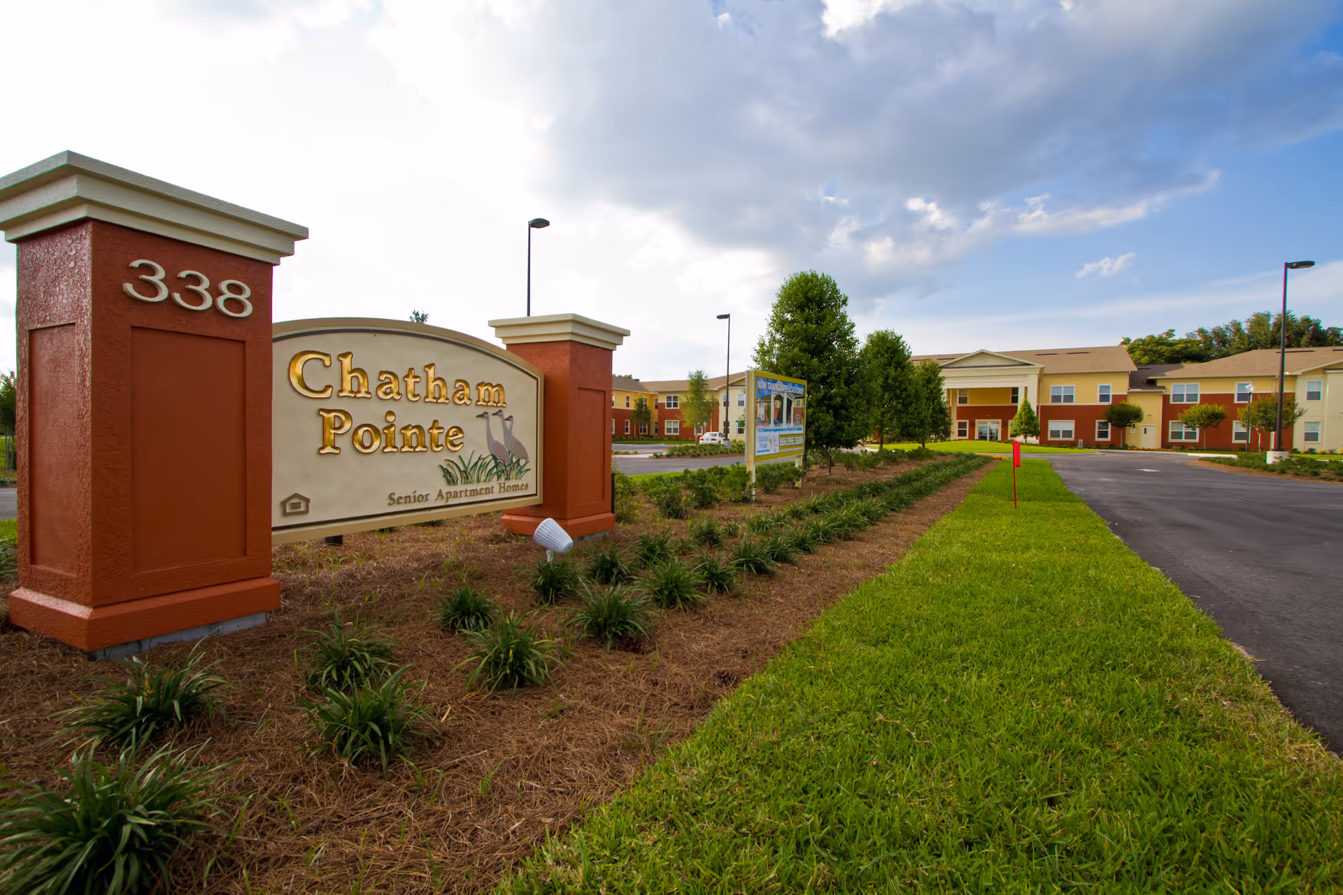 Entrance sign for Chatham Pointe Senior Apartment Homes with the address number 338 on a brick pillar, surrounded by landscaped greenery and a paved driveway leading to a two-story building under a partly cloudy sky.