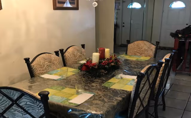A dining table set for four with patterned placemats, napkins, and glasses. The table has a decorative centerpiece with candles and red flowers. The room has tiled flooring and a door with glass panels in the background.