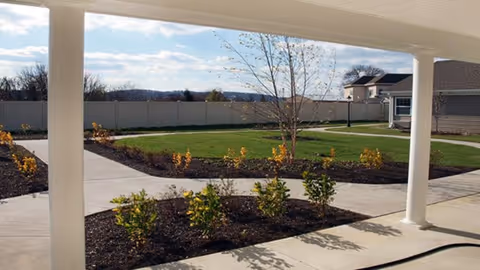 Covered walkway overlooking a landscaped courtyard with lawn, shrubs, a young tree, and a surrounding fence.