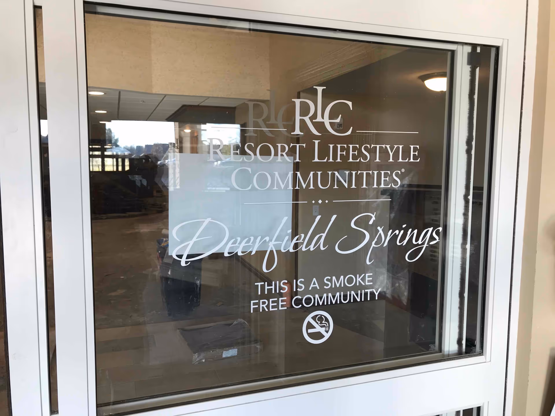 Glass entrance window with white vinyl lettering reading 'Resort Lifestyle Communities Deerfield Springs' and 'This is a smoke free community' with lobby reflections visible.