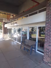 Exterior view of a building entrance with a covered walkway. There is a bench placed on the sidewalk in front of large glass windows and doors. The building has a brick facade and a white sign above the entrance with the text 'Fairhaven'. Trees and parked cars are visible in the reflection on the glass.