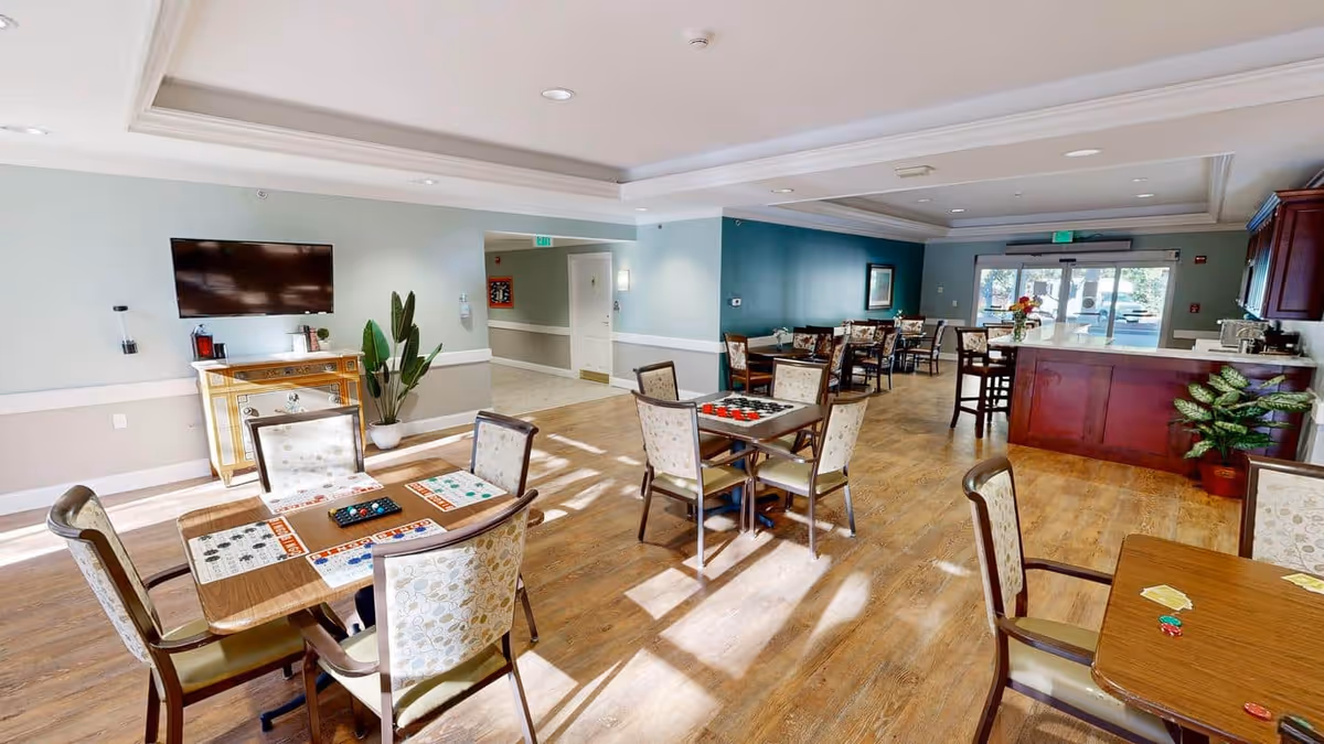 A bright and spacious common area in a senior living facility with multiple tables and chairs arranged for games and social activities. The room features wood flooring, a mounted flat-screen TV, a decorative cabinet, potted plants, and a counter area with additional seating near the entrance.