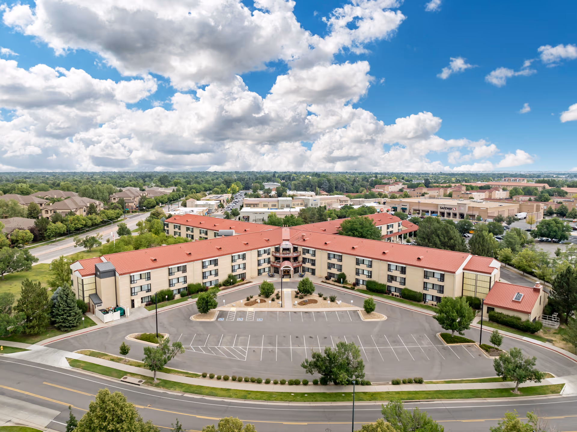 Aerial view of Brookdale Lowry, a senior living facility with a large U-shaped building featuring a red roof and beige walls. The building is surrounded by trees, parking spaces, and roads. In the background, there are other buildings, a Safeway store, and a partly cloudy blue sky.