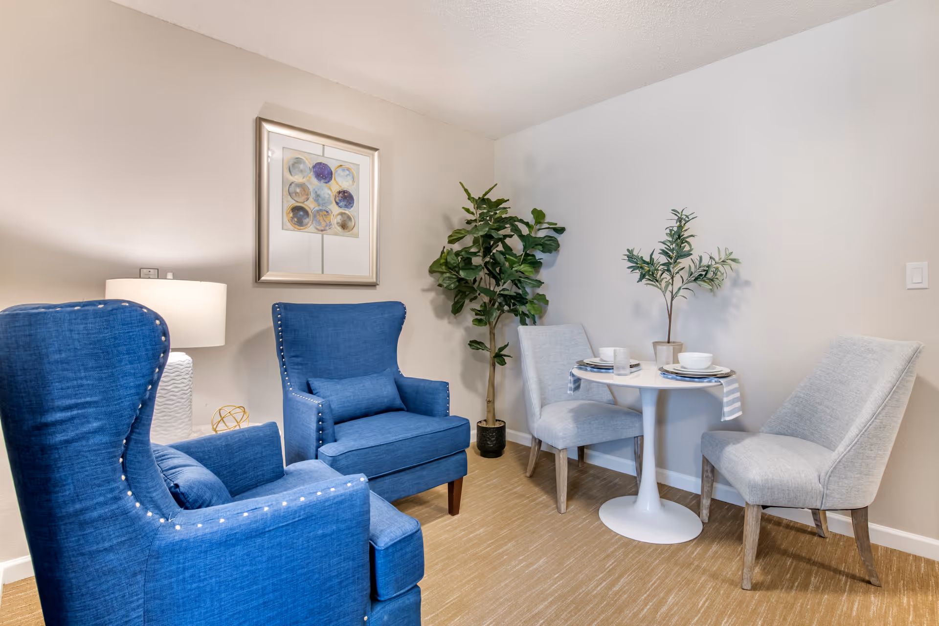 Small sitting area with two blue armchairs, a round white bistro table with two gray chairs, and plants and wall art.