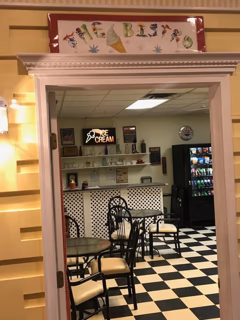 View through a doorway into a small bistro area with black and white checkered floor, several black metal chairs with cream cushions around glass-top tables, a counter with shelves holding various items, a vending machine, and a neon sign that says 'ICE CREAM'. Above the doorway is a colorful sign that reads 'THE BISTRO' with an ice cream cone illustration.