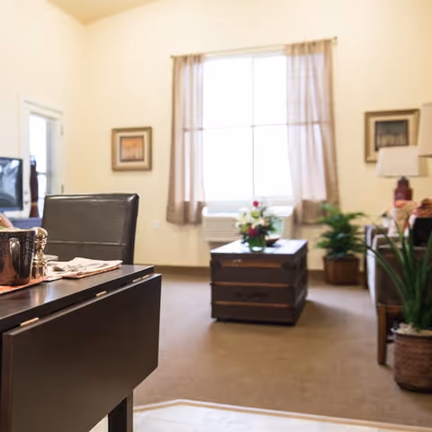 Bright living room featuring a trunk-style coffee table topped with flowers, seating, plants, and a curtained window.