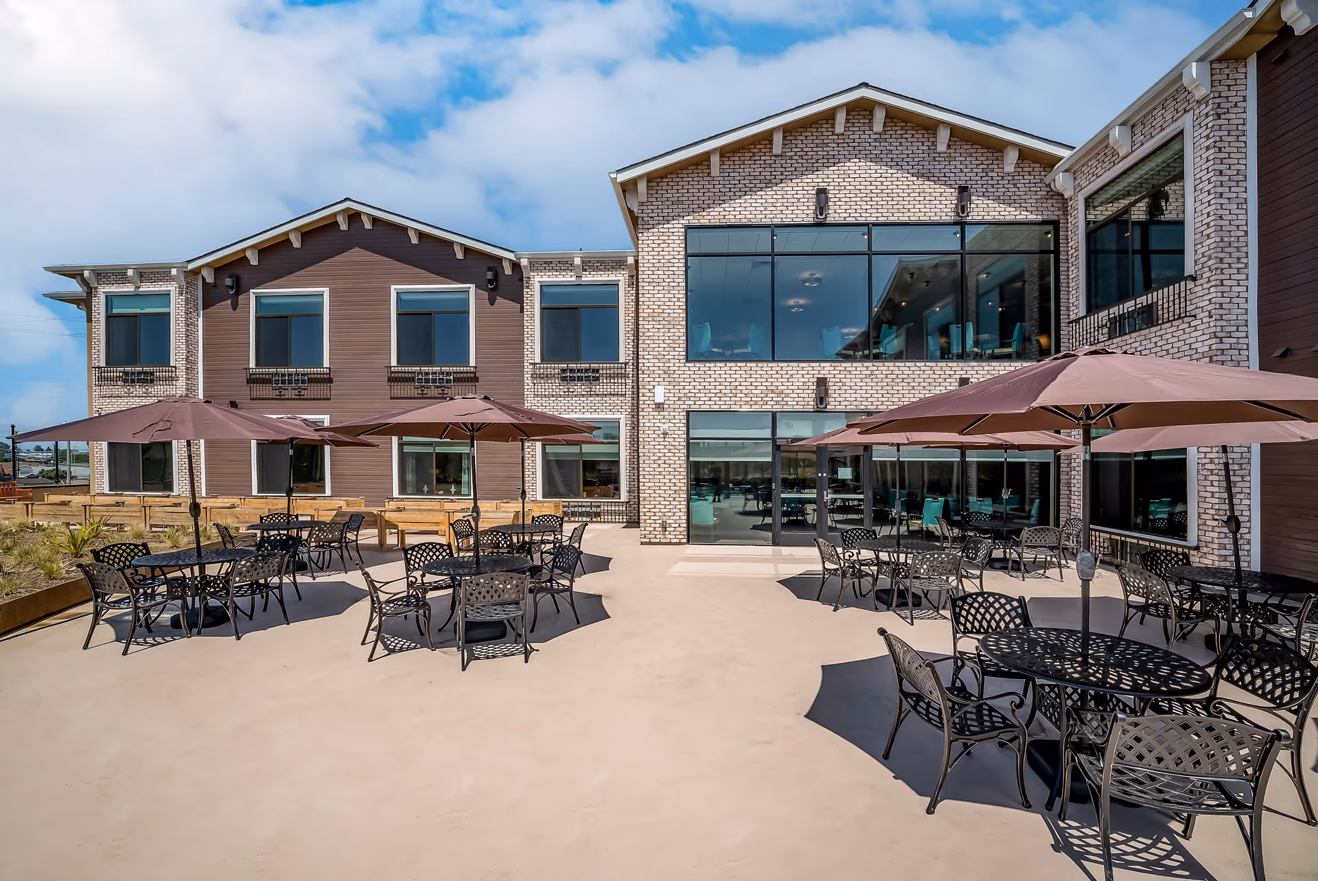 Outdoor patio area at Beach Terrace with several black metal tables and chairs, each shaded by large brown umbrellas. The patio is adjacent to a two-story building with a combination of brown siding and light brick exterior. Large windows on the building reflect the outdoor seating area and provide views into the interior.
