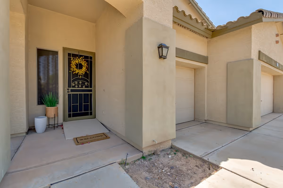 Exterior view of a senior living facility entrance with a green door decorated with a yellow wreath, a welcome mat, two potted plants, and adjacent garage doors under a beige stucco archway.