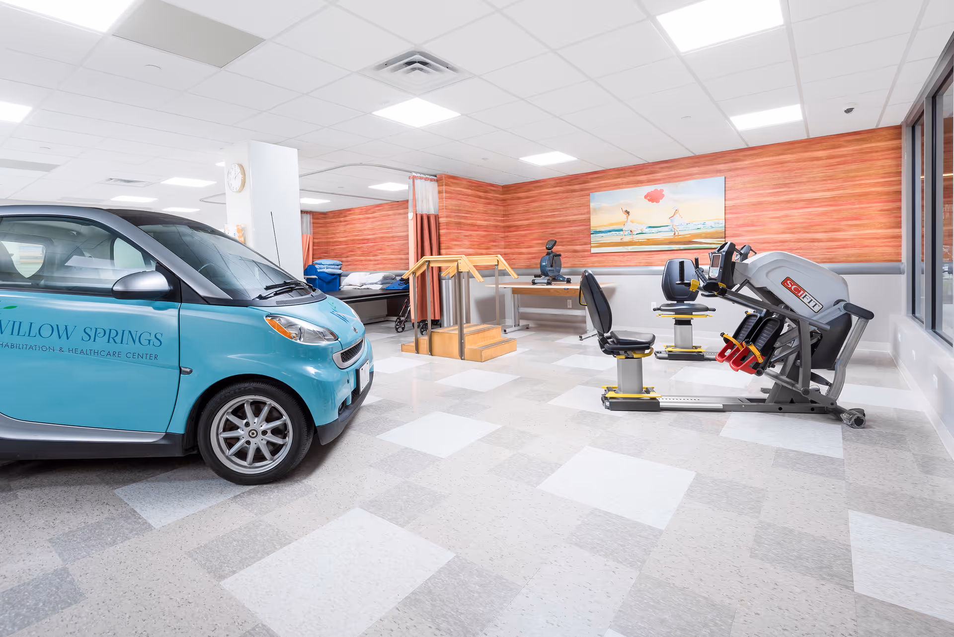Interior room of Willow Springs Rehabilitation & Healthcare Center featuring a small blue car with the facility's name on it, exercise equipment including a seated leg press machine, parallel bars for physical therapy, a hospital bed, and a colorful painting on the wall.