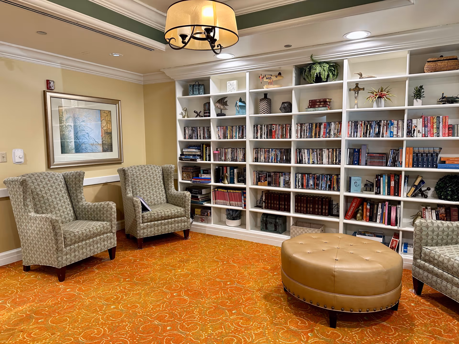 A cozy reading room with two patterned armchairs, a large round tan ottoman, and a white built-in bookshelf filled with books, DVDs, and decorative items. The room has warm yellow walls, an orange patterned carpet, a framed artwork on the wall, and a ceiling light fixture.
