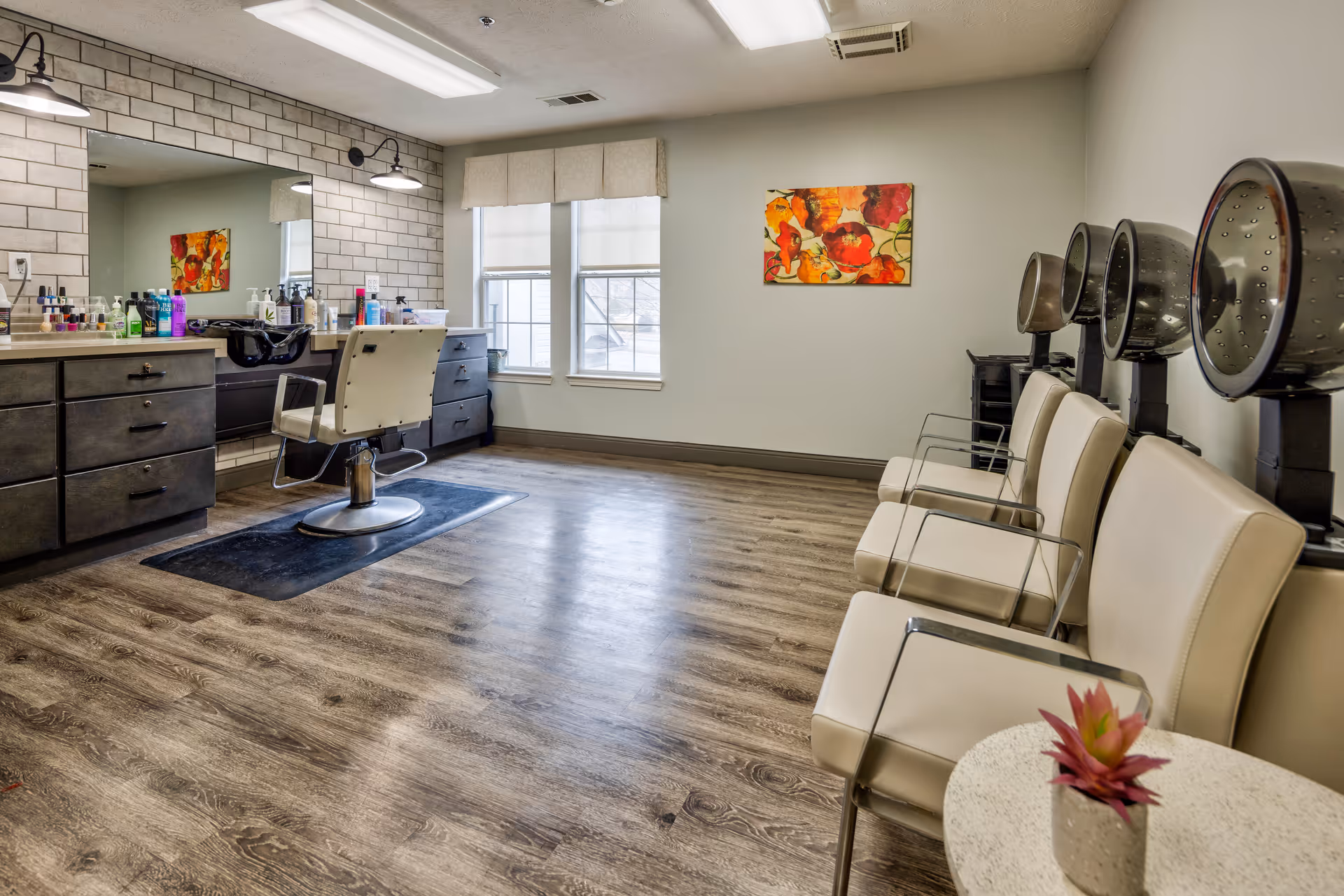 Interior of a hair salon area with a styling chair in front of a large mirror, hair care products on the counter, three hair drying stations with chairs along the right wall, a small round table with a potted plant, and a colorful floral painting on the far wall under two windows.