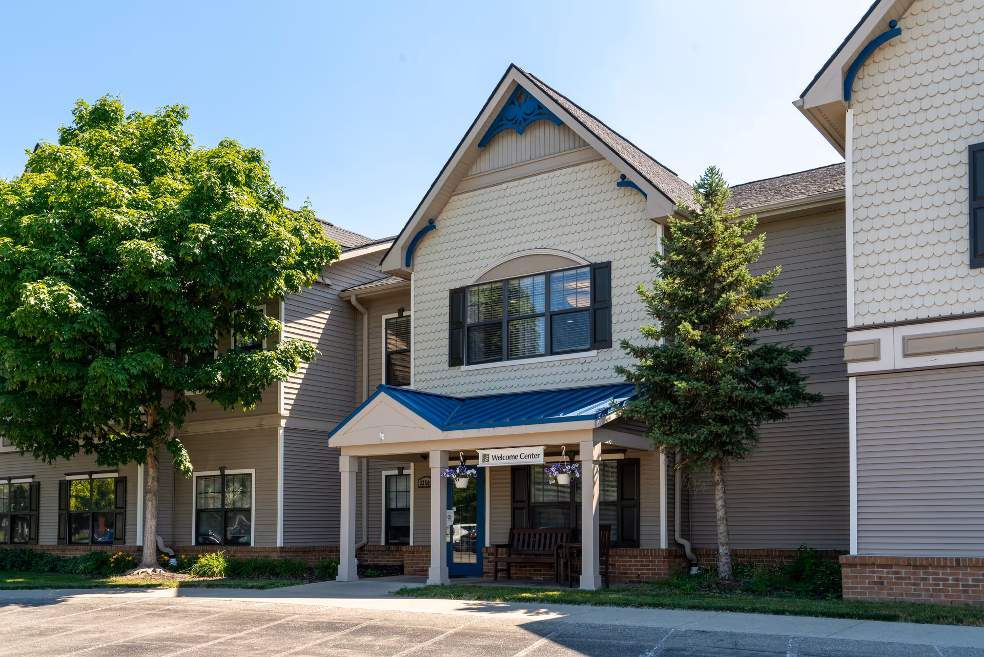 Exterior view of Commonwealth Senior Living at New Baltimore showing the entrance to the Welcome Center with a covered porch, two hanging flower pots, a bench, and surrounding trees under a clear blue sky.